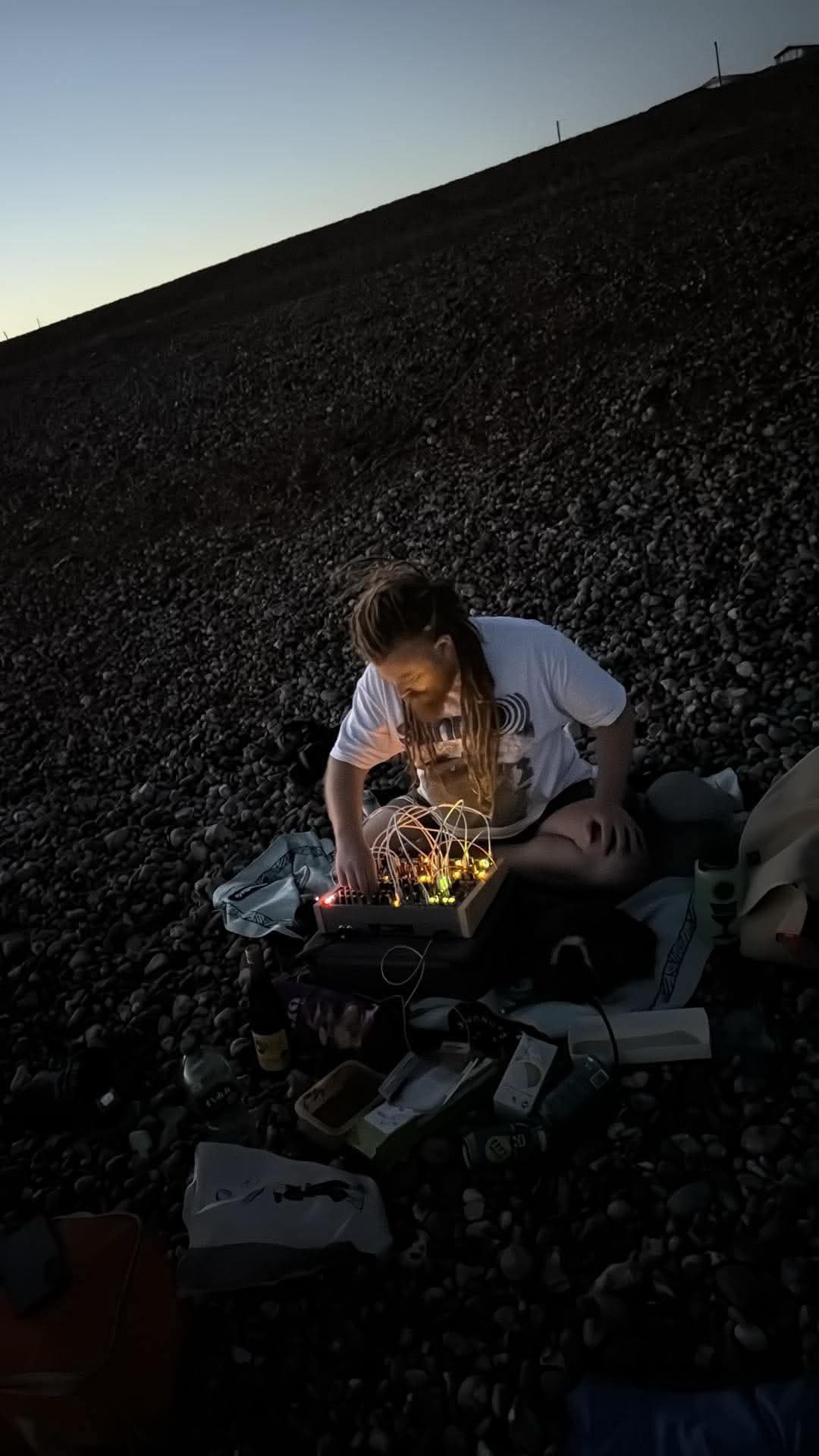 A wizard sat cross legged on a pebble beach playing with an analog synthesiser