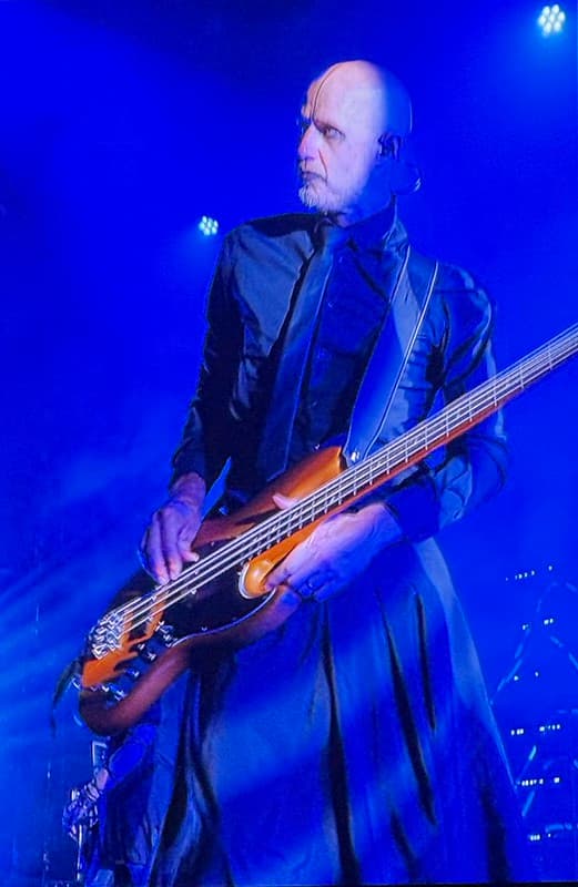 Bass player, with blue light in the background, he has a bald head and a long black costume
