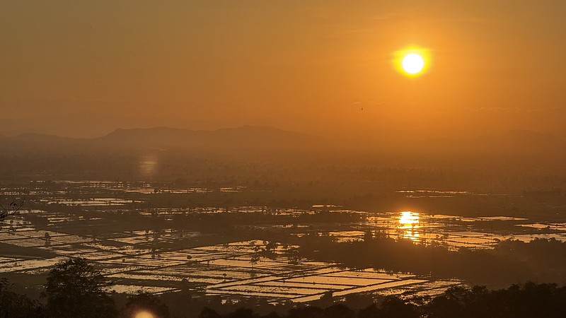 The sunset taken from the top of a hill, in the distance you can see some hills. The sunset is orange and can be seen reflected in the rice fields below