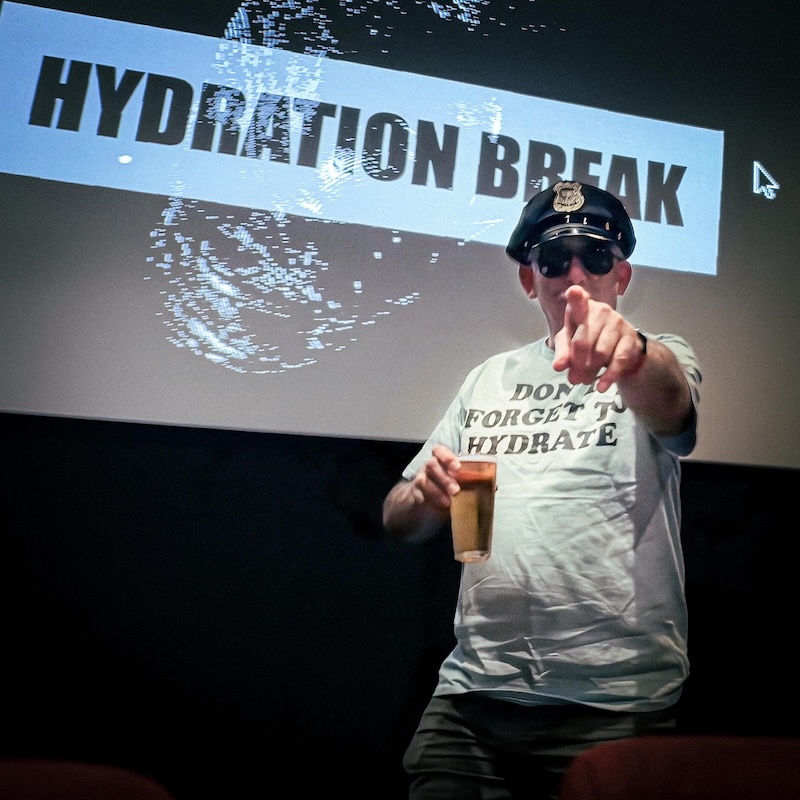 Me dress as a Police Officer wearing a t-shirt that reads: Don't forget to hydrate, Holding a a drink and pointing (photo by Denis Frank)