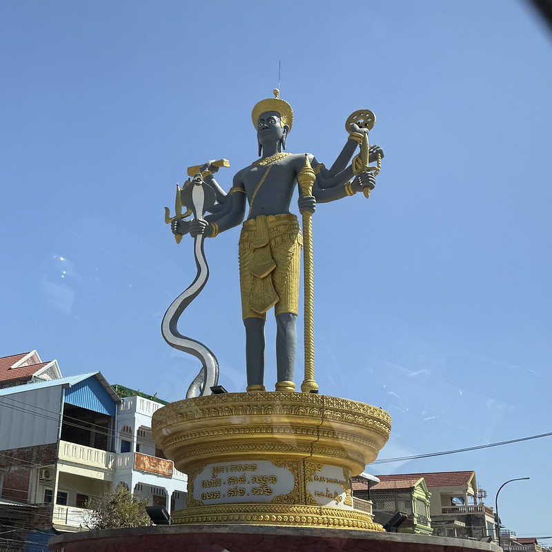 A roundabout in Battambang, it has a circular gold pedestal, stood atop is a black man with 8 arms who is wearing gold shorts. In one of his right arms he is holding a snake and in one of his left arms a gold staff
