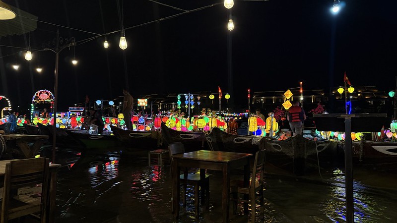 The view from our table at the restaurant, in the foreground is a table and chairs stood in the water behind them are a number of boats all lit up with colourful lanterns