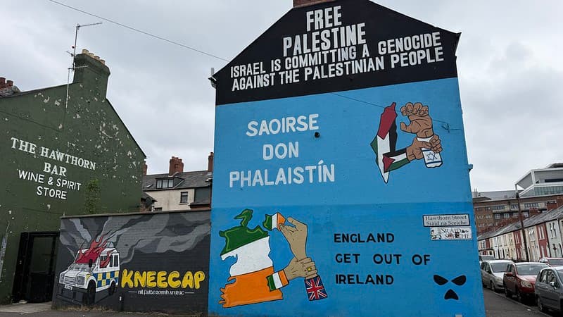 The Kneecap Mural in West Belfast, on the left there is a police vehicle on fire. On the right is a map of Ireland with a hand grabbing back Northern Ireland back from Britain. At the top it reads: Free Palestine Isreal is committing  a genocide against the  Palestinian people