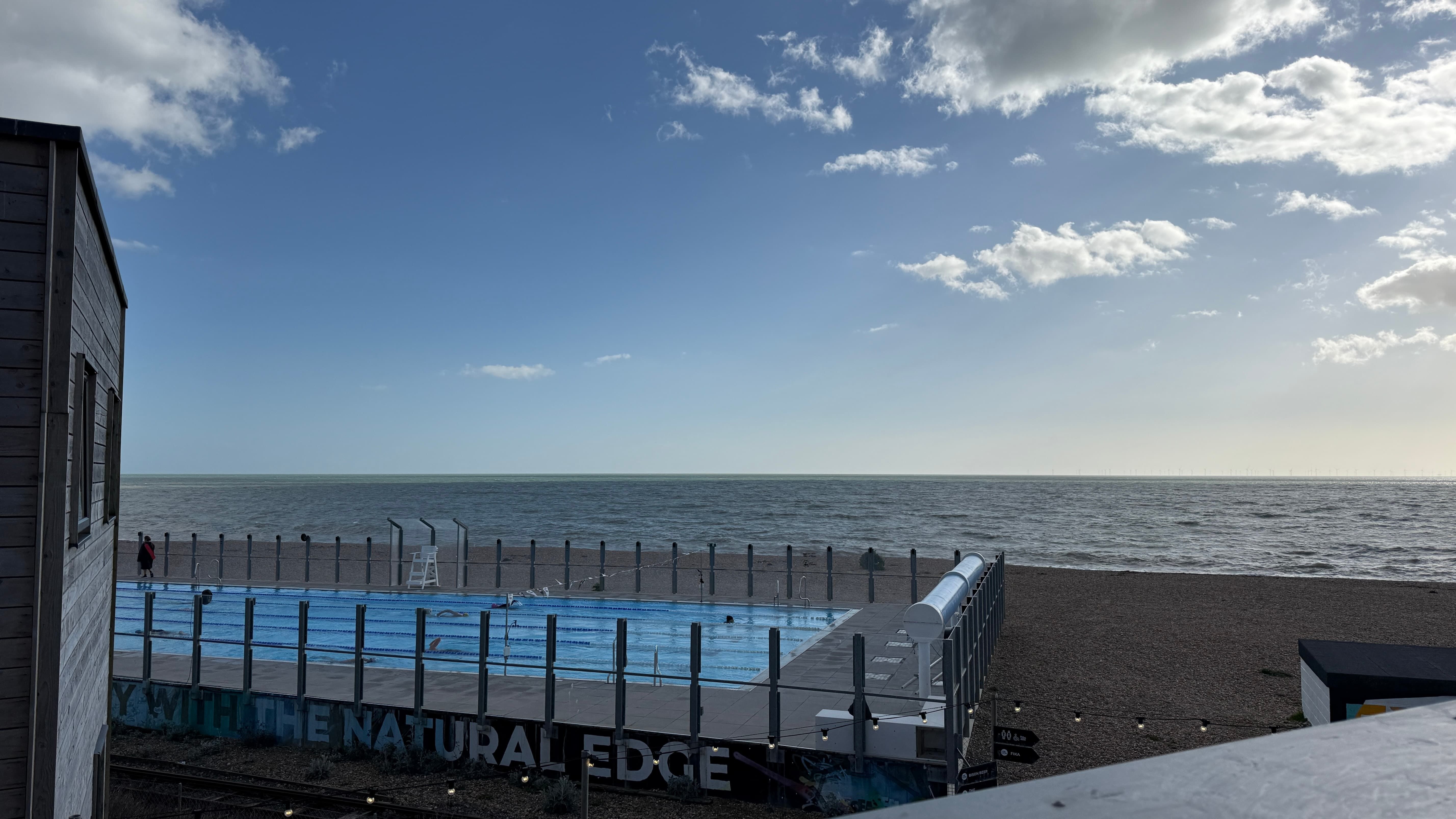 View from the roof of the Bison Beach Bar, there is a swimming pool with lanes on a pebble covered beach the sky is mostly blue with a few fluffy clouds